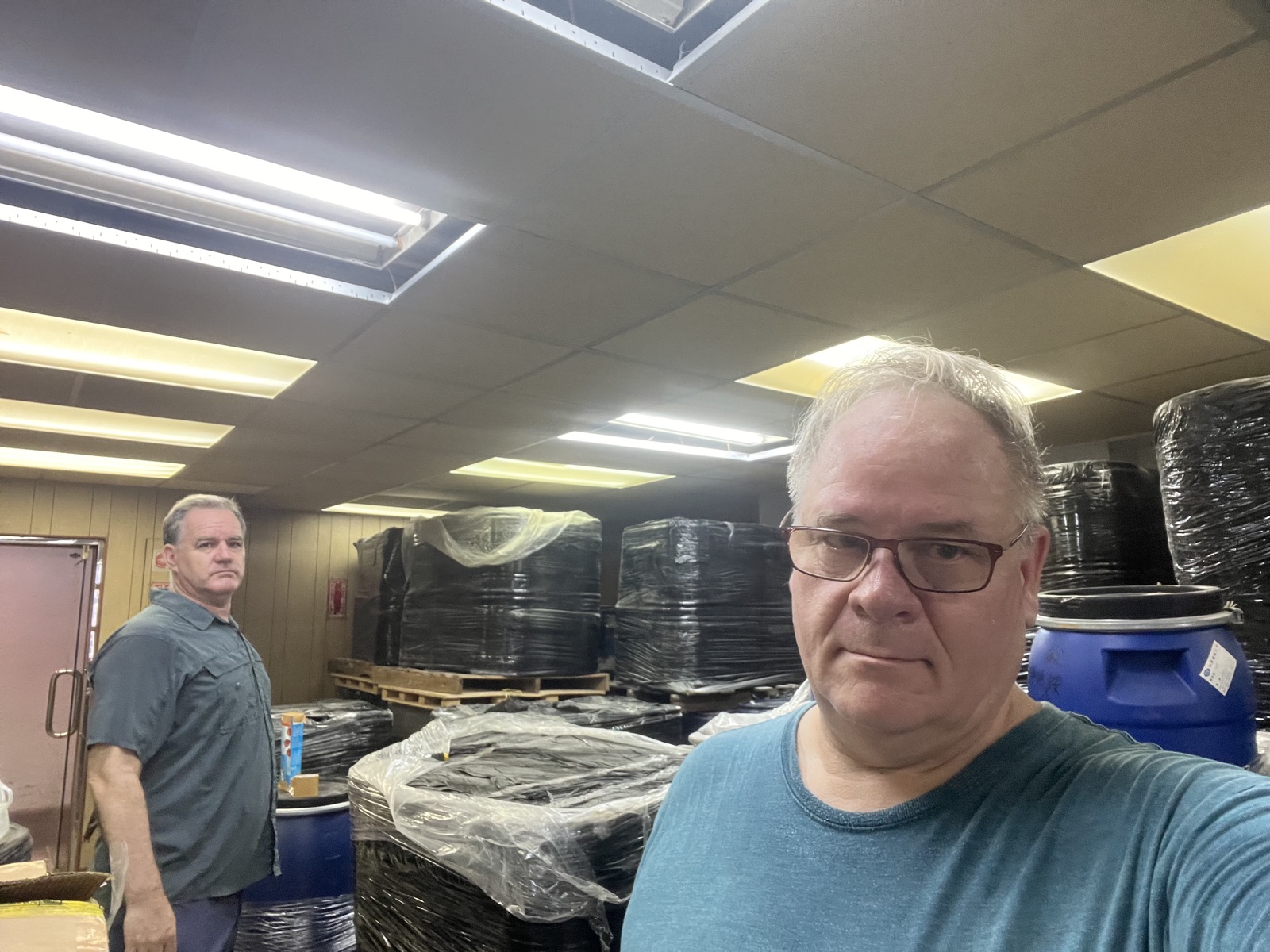 Two people standing in a warehouse among stacked, wrapped storage drums